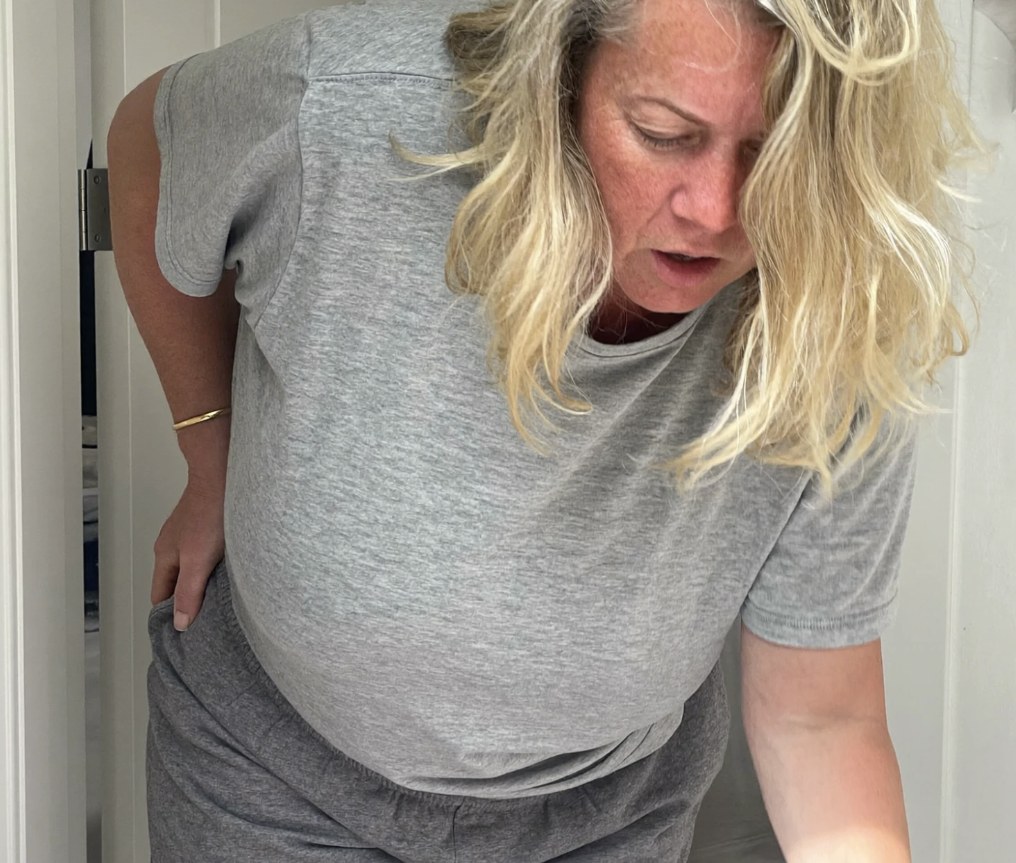 A woman with blonde hair bent over in a doorway, hand pressed to her lower back, stiff and aching