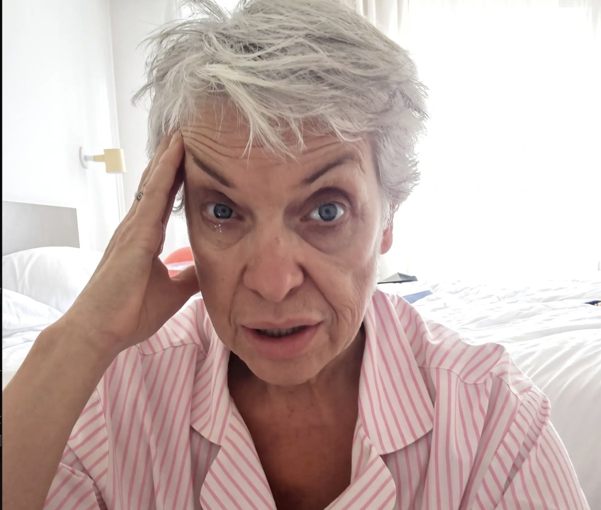 A woman with short grey hair sitting on her bed in striped pyjamas, hand pressed to her temple, looking exhausted and foggy